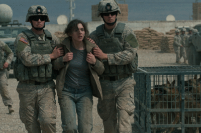 “They Hurled Her Toward Starving K9s While Yelling ‘Get Destroyed’ — Never Realizing She Owned Every One of Them”  “Get that civilian pencil pusher out of my kennel. This is a war zone, not a petting zoo. Someone remove her before she gets one of my men killed.”  The words cracked through the heat like a whip.  Around Captain Valyrias, a loose semicircle of security forces shifted their weight and let out a thin, uneasy snicker. Not laughter. Not really. The kind of sound people make when they sense something ugly unfolding and decide it’s safer not to notice.  Bram Air Base baked under the sun. Heat shimmered off concrete and blast walls, turning every breath into effort. The K9 compound echoed with restless barking—deep, hungry, territorial. These weren’t ceremonial dogs. They were lean, sharp-eyed, and deliberately kept on edge.  Valyrias stood with his hands planted on his hips, compact and dense, built like a bulldog that had never learned restraint. His neck nearly merged with his shoulders, his stance broadcasting one message: this is mine.  With a sharp flick of his chin, he pointed toward the woman standing at the compound gate.  She didn’t look like she belonged.  Average height. Slight build. Hair pulled back into a tight, functional knot. Plain fatigues—no rank, no name tape, no insignia. Nothing that fit neatly into the hierarchy Valyrias worshiped. Just one small circular patch on her shoulder: a wolf’s head framed by a shepherd’s crook. Obscure. Unfamiliar. Easy to dismiss.  She didn’t flinch.  She didn’t blink.  As Valyrias barked orders and gestured toward the kennels, she remained still—calm, centered, unreadable. Her gray eyes moved slowly across the compound, cataloging everything: the dogs’ posture, the handlers’ grip on the leads, the tension threaded through the air. When her gaze finally settled on Valyrias, it wasn’t defiant.  It was analytical.  He mistook that silence for weakness. For fear. For confirmation that he was in control.  “Get her closer,” someone muttered. “Let her see why she doesn’t belong here.”  The dogs lunged against their restraints, muscles coiling, teeth flashing. A few men laughed louder now, adrenaline feeding their cruelty.  Two hundred yards away, in an office cooled by humming air vents, Colonel Madson stared at a grainy security monitor. The image was warped by distance and heat distortion—but one detail cut through it all.  The way she stood.  Balanced. Relaxed. Ready.  Madson felt his stomach drop.  He hadn’t seen that posture since his years embedded with the most elite special operations units—the kind of people who didn’t need to announce authority because they were authority.  His hand hovered over the phone.  Because in that moment, Colonel Madson knew something no one in the kennel realized yet:  Captain Valyrias wasn’t asserting dominance.  He was humiliating himself in front of the one person on that base who didn’t answer to him at all.  And when the truth surfaced—about who she was, why she was there, and who those K9s truly belonged to—  this wasn’t going to end with barking.  (The story continues in the first comment.)